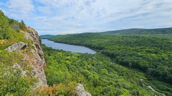 View from a rocky escarpment in Michigan's Porcupine Mountains across Lake of the Clouds and unbroken green forest to rolling hills under a bright, partly cloudy sky.